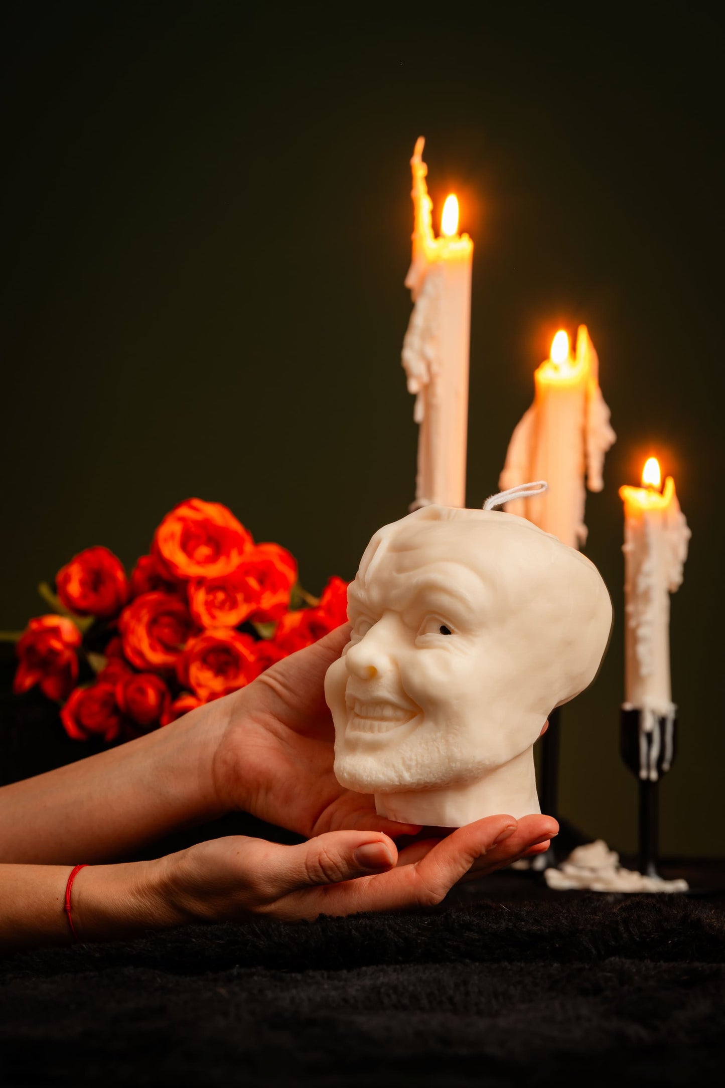 White ceramic head sculpture held by a hand with lit candles and red flowers in the background