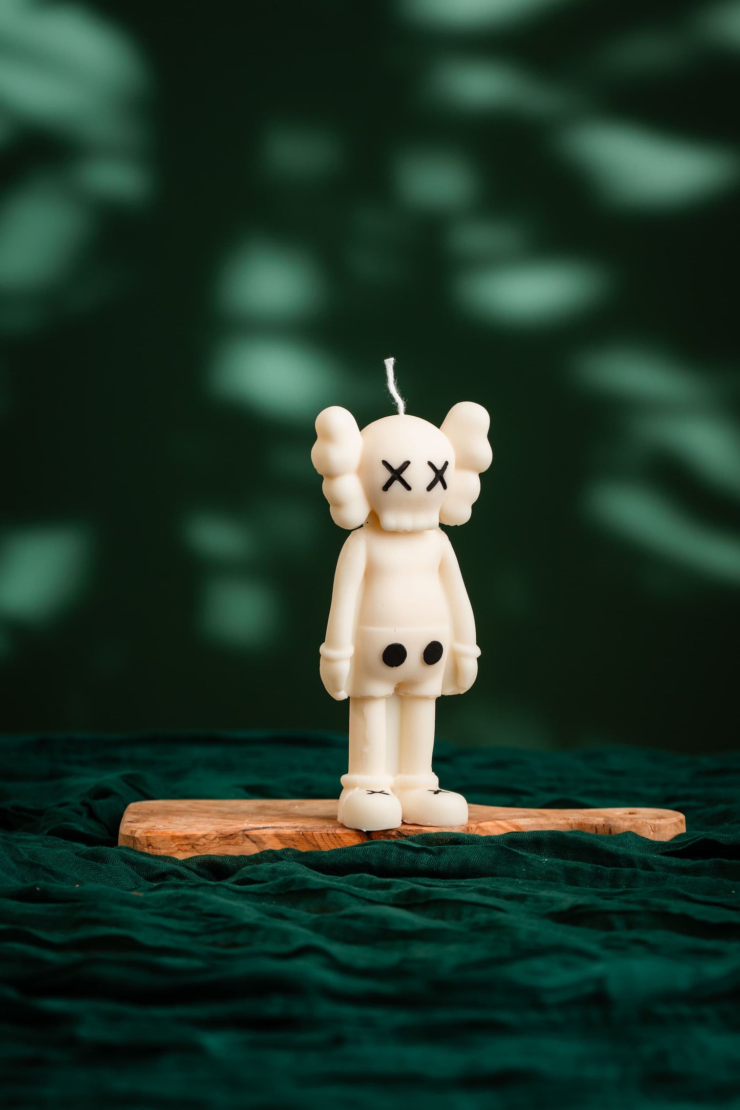 White bear-shaped candle with black eyes on a wooden stand against a dark green background, KAWS Inspired Standing Candle – Vegan & Non-Toxic