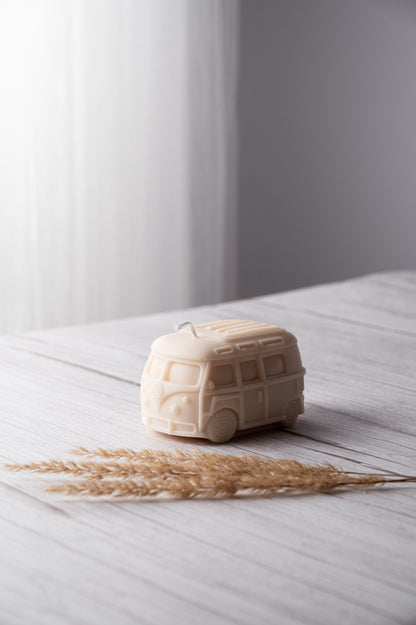 A beige wax candle shaped like a retro bus placed on a wooden surface with some grain, with a decorative dried plant beside it.