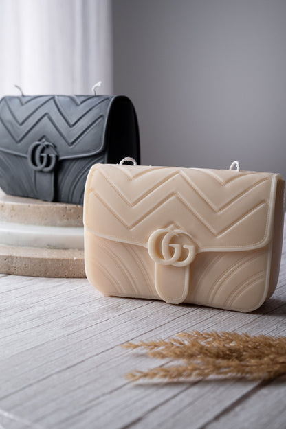 Two soy wax candles designed to resemble luxury handbags in beige and black colors, placed on a wooden surface with a decorative element in the background.