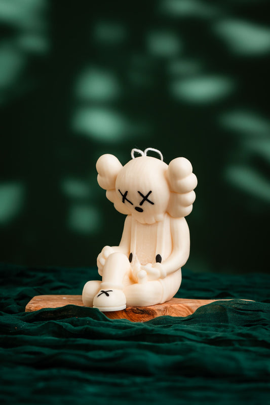 White plush toy with black accents on a wooden platform against a dark green background, KAWS Passing Through Game Over Candle – Handmade Soy Wax