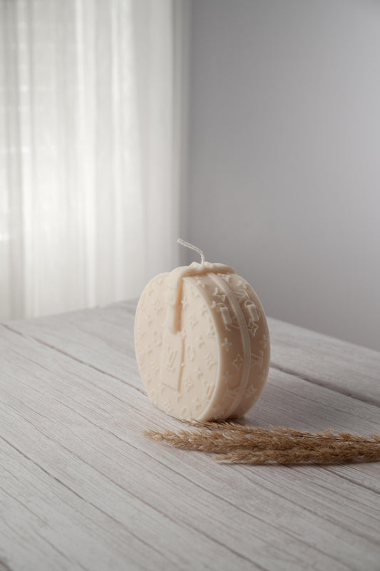 A beige, abstract-patterned candle on a white surface with a shadow falling over part of the image, indicating a warm light source.
