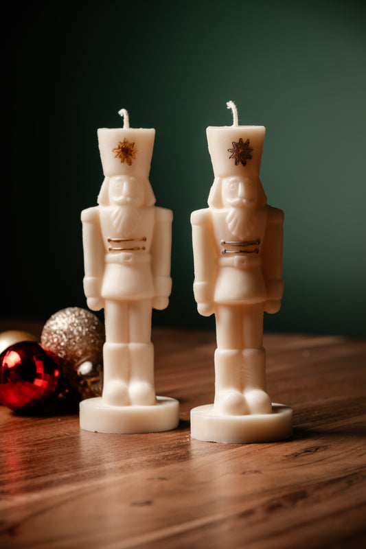 Two soy wax candles shaped like nutcrackers, placed on a wooden surface with a green background and decorative elements around.