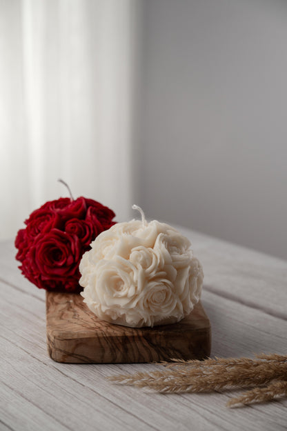 A candle with a rose flower ball design on top, placed on a wooden board, with a red rose and white candle wax in the background.