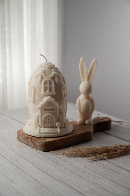 A beige candle in the shape of a house with a bunny figure, placed on a wooden board.