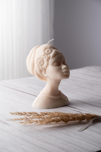 A beige soy wax candle shaped like an African female figure with a decorative headpiece, placed on a wooden surface with dried plant reeds to the side.